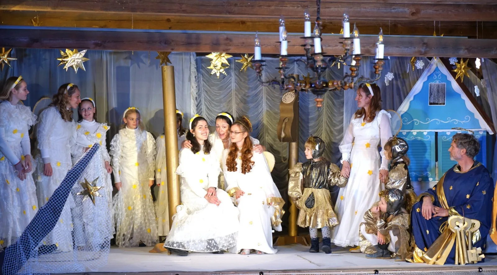 Engelgesellschaft im Himmel mit Sternenglanz – Weiße Engel mit goldenen Sternen stehen und sitzen im Himmelszelt, Kinder in goldenen Kleidern und der Hüter mit großen Schlüsseln rahmen ein blaues Häuschen – feierliche, warme Ensemble-Szene.