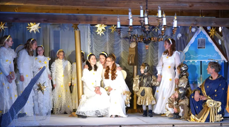 Engelgesellschaft im Himmel mit Sternenglanz – Weiße Engel mit goldenen Sternen stehen und sitzen im Himmelszelt, Kinder in goldenen Kleidern und der Hüter mit großen Schlüsseln rahmen ein blaues Häuschen – feierliche, warme Ensemble-Szene.