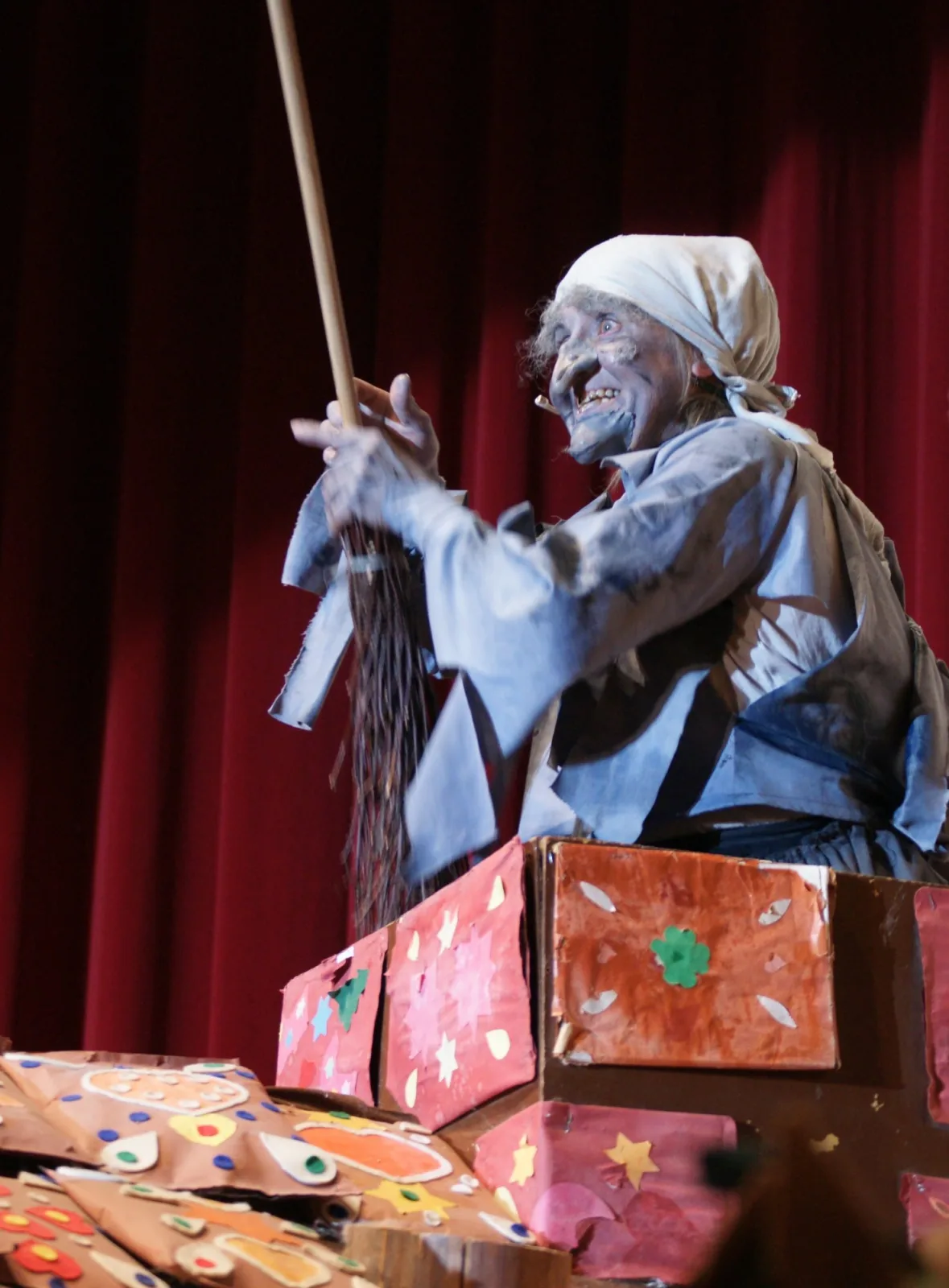 Hexe im Schornstein auf dem Knusperhäuschen mit erhobenem Besen – Die grau geschminkte Hexe mit Kopftuch schwingt lachend den Reisigbesen über bunten Lebkuchen auf dem Dach des Knusperhäuschens – frecher Auftritt mit Augenzwinkern.