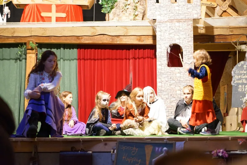 Kinder auf der Bühne beim Halloween-Kürbisfest – Kinder in bunten Kostümen spielen Szenen des Märchen-Musicals „Nächtliche Bisse“ auf der Bühne, mit Vampir- und Hexenmasken vor Burgkulisse.