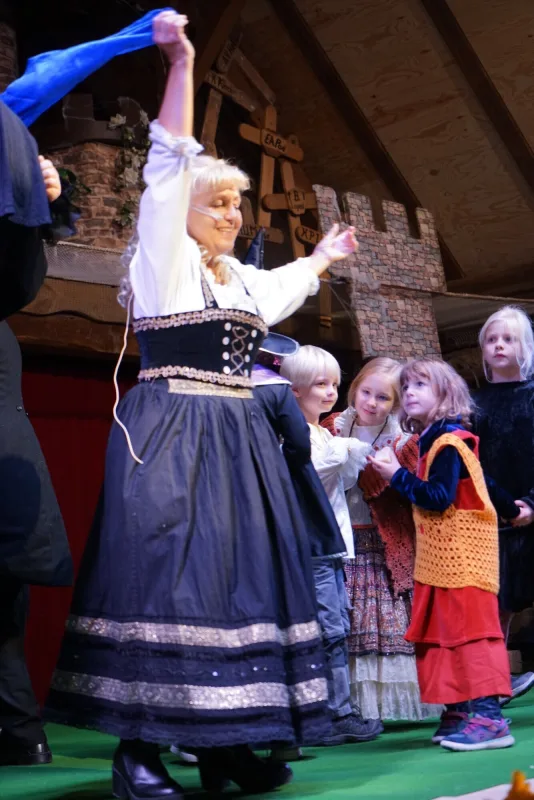 Bäckerin mit Kindern auf der Musicalbühne – Die Bäckerin hebt freudig ein Tuch, umgeben von Kindern in Kostümen, während die Szene des Dorfes lebendig wird.