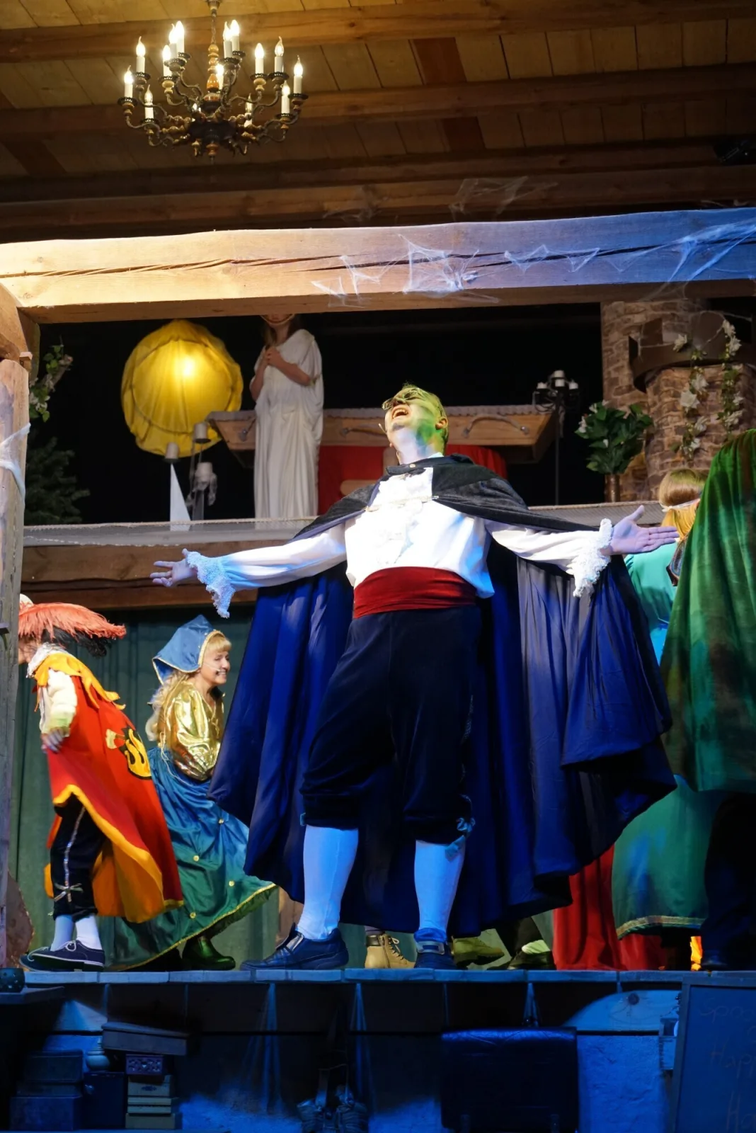Graf Dracula triumphiert im Schlossgarten – Graf Dracula mit ausgebreiteten Armen und dunklem Umhang genießt die Aufmerksamkeit im Bühnenlicht, umgeben von maskierten Figuren.