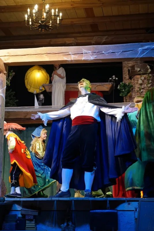 Graf Dracula triumphiert im Schlossgarten – Graf Dracula mit ausgebreiteten Armen und dunklem Umhang genießt die Aufmerksamkeit im Bühnenlicht, umgeben von maskierten Figuren.