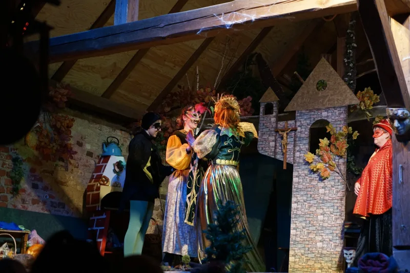Begegnung am Maskenball in der Schlosskulisse – Maskierte Darsteller in goldenen und roten Kostümen interagieren lebhaft auf der Bühne vor der Burgwand.