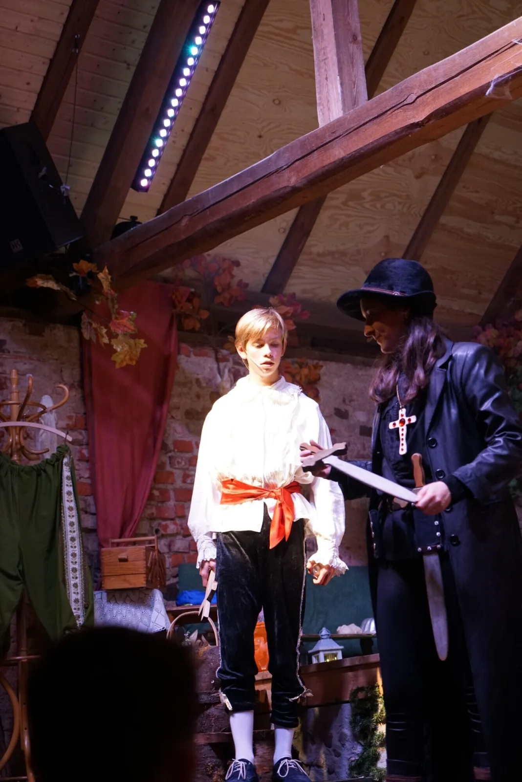 Julia und Conan mit Kreuzen bereit zum Kampf – Julia im schwarzen Ledermantel und Conan mit rotem Gürtel stehen bewaffnet mit Kreuzen und Schwertern auf der Bühne.