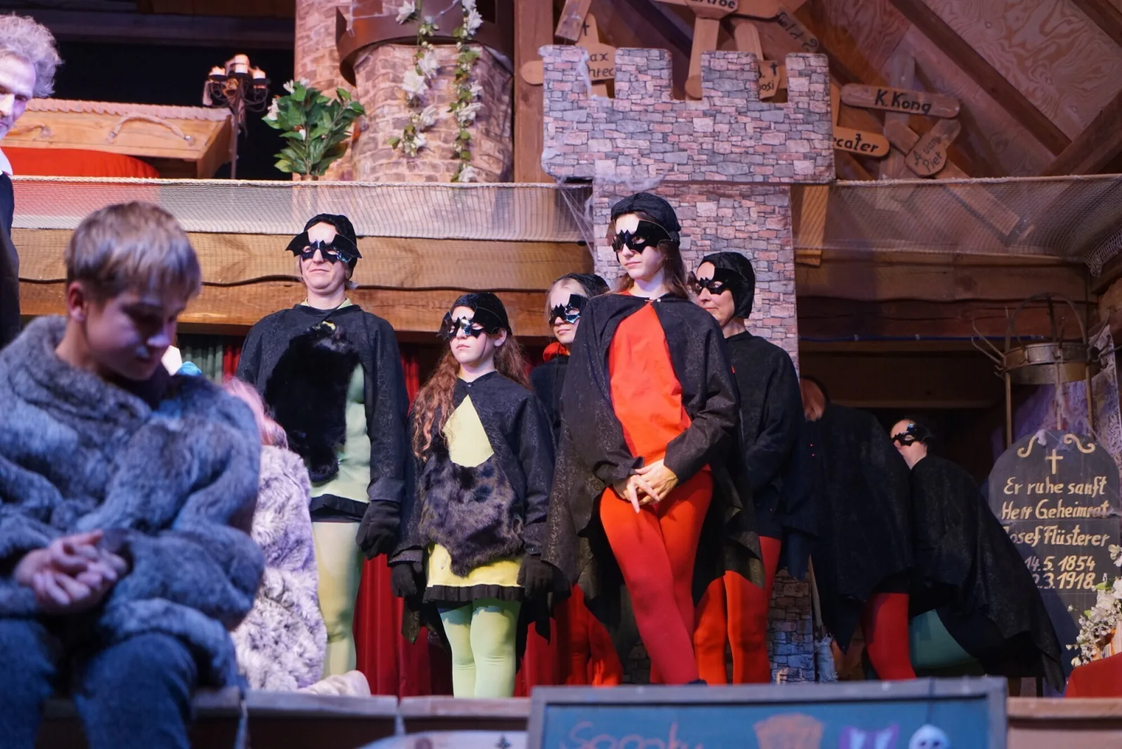 Fledermausensemble beim Finale in Schlosskulisse – Mehrere Darsteller im Fledermauskostüm mit roten und grünen Akzenten stehen beim Schlussbild der Aufführung auf der Bühne.